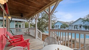 A covered outdoor area with a wooden deck, red Adirondack chairs, a metal table, and a view of a pool and buildings in the background. - 2 BR Cottage in 30A | Outdoor Pool, 2 Bikes | My Beach Getaways (Inlet Beach)