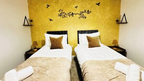 Et hotelværelse med to senge, hver med en hvid madras og et beige tæppe. Der er vægmonterede lamper på hver side af sengene, og en dekorativ vægkunst med fugle og grene.