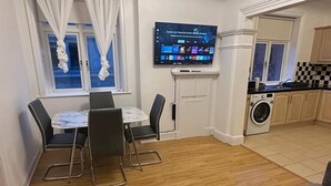 Een moderne woonkamer met een grijze bank, een eethoek met een tafel en stoelen, een aan de muur bevestigde televisie, en een keuken met houten kastjes en een wasmachine.