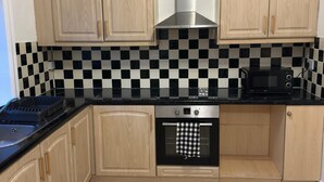Een keuken met licht houten kastjes, een zwart-wit geruit spatwand, een roestvrijstalen afzuigkap, een zwarte magnetron en een betegelde vloer.