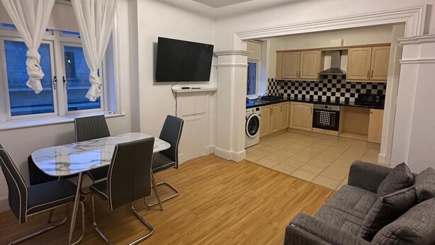 Een moderne woonkamer met een eethoek, een grijze bank, een flatscreen televisie aan de muur en een keuken op de achtergrond.