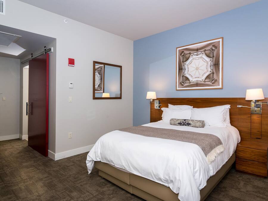 A hotel room with a bed, two bedside lamps, a wall-mounted artwork, a door, and a red exit sign.