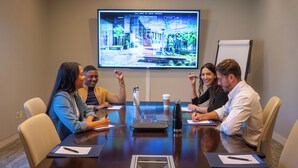 Un salón de conferencias con una pantalla grande mostrando una imagen de un edificio, una pizarra y una mesa con computadoras portátiles, papeles y tazas de café.