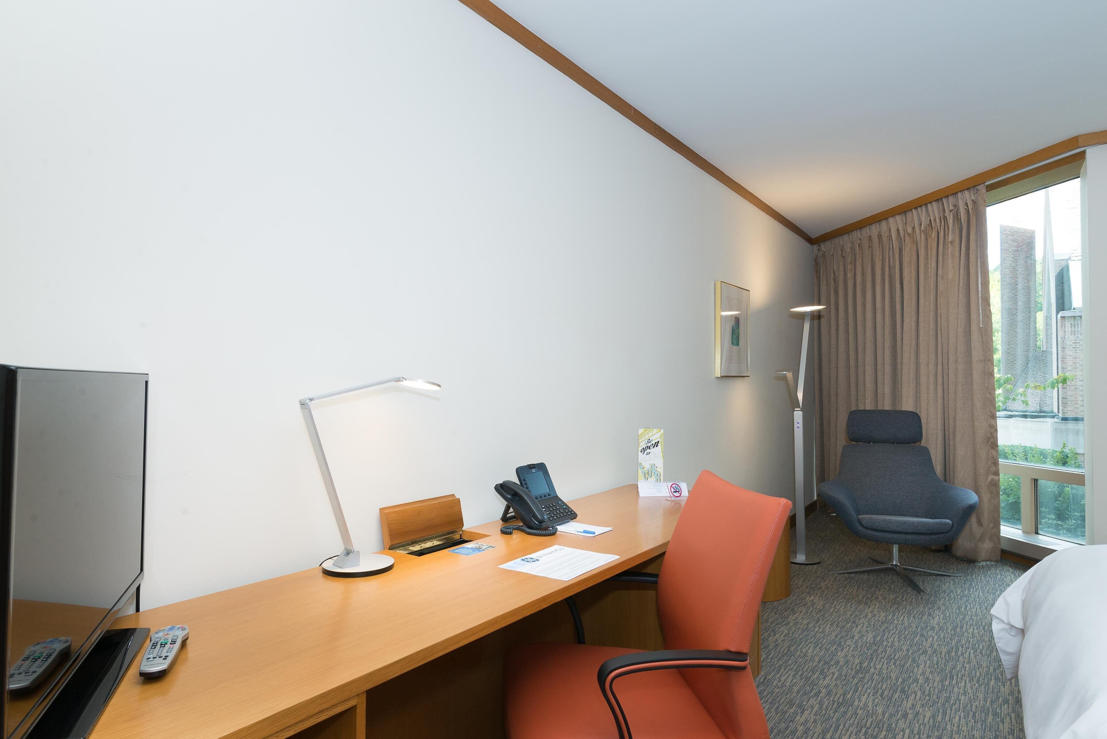 Hotelkamer met bureau, stoel, bed, televisie en raam met gordijnen.