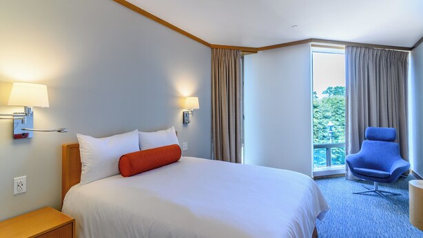 Una habitación de hotel con una cama grande, dos lámparas de noche, una mesita de noche, un sillón azul y vistas a zonas verdes a través de la ventana.