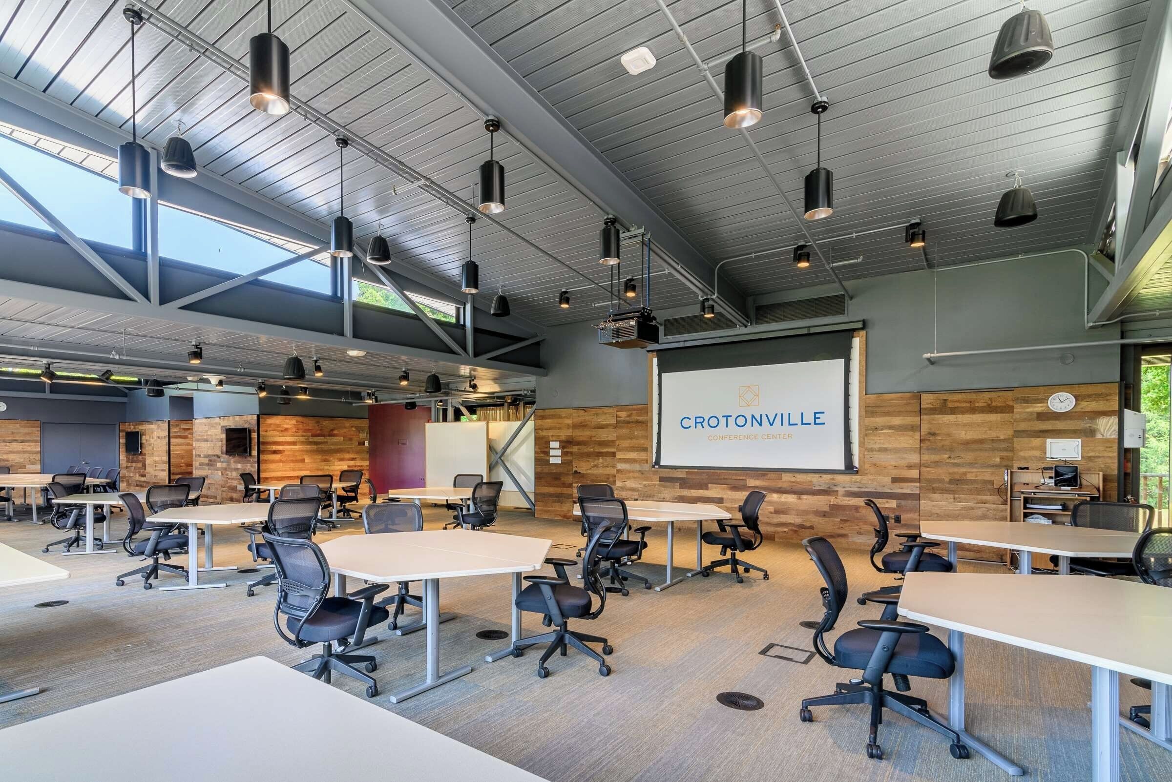 A conference room with multiple tables and chairs, a large screen displaying 'CROTONVILLE CONFERENCE CENTER', and a high ceiling with industrial-style lighting.