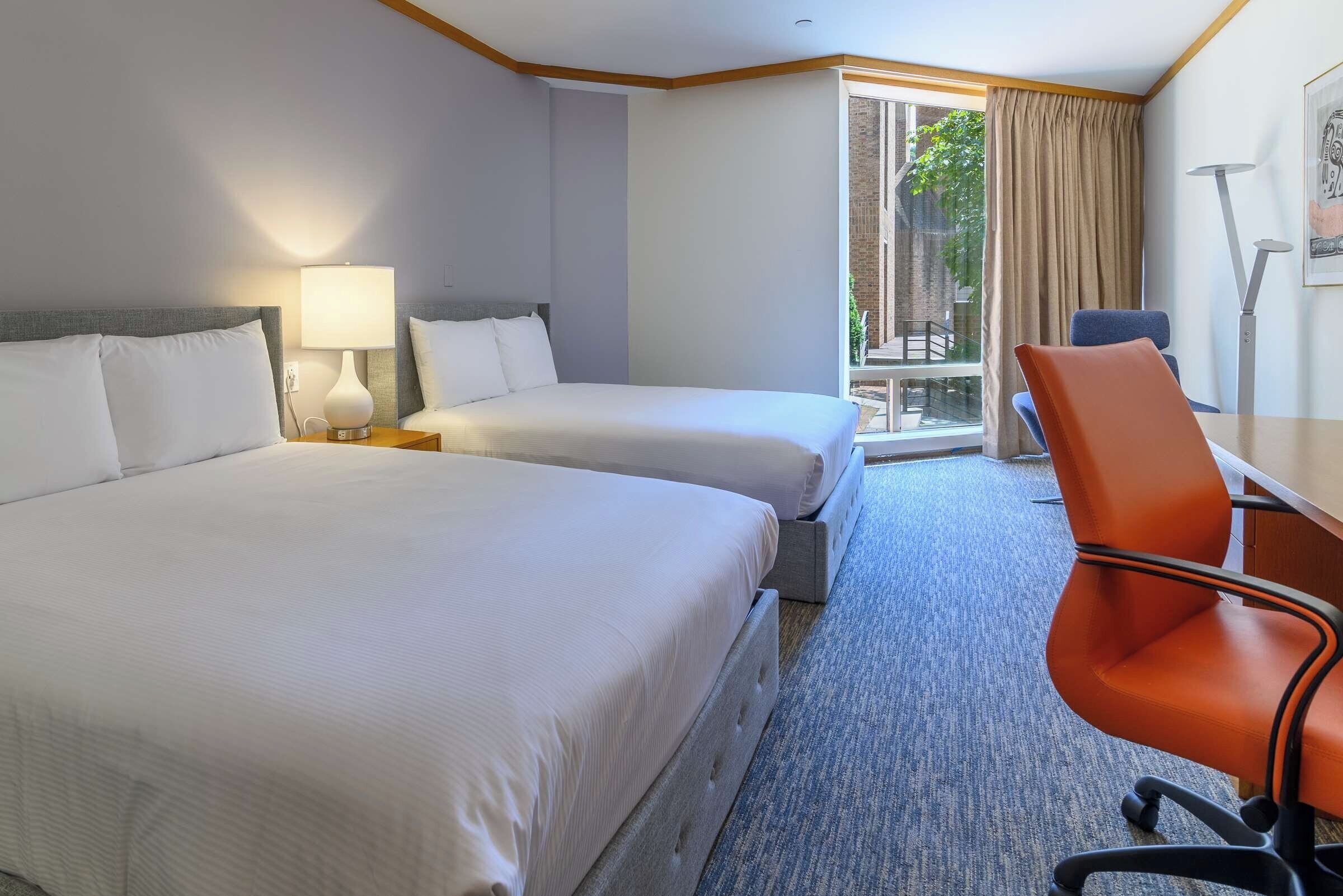 Habitación de hotel con dos camas, un escritorio, una silla y una ventana con vistas al exterior.
