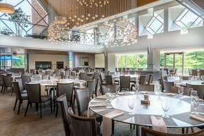 A spacious dining area with multiple tables and chairs, large windows, and a modern chandelier. - Windrose On Hudson (Ossining)
