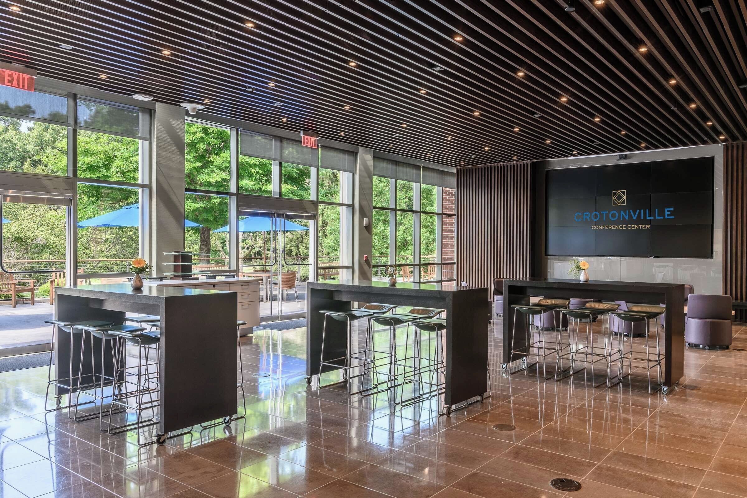 Un moderno centro de conferencias con grandes ventanales, una zona de bar y una pantalla de proyección que muestra el nombre 'CROTONVILLE CONFERENCE CENTER'.