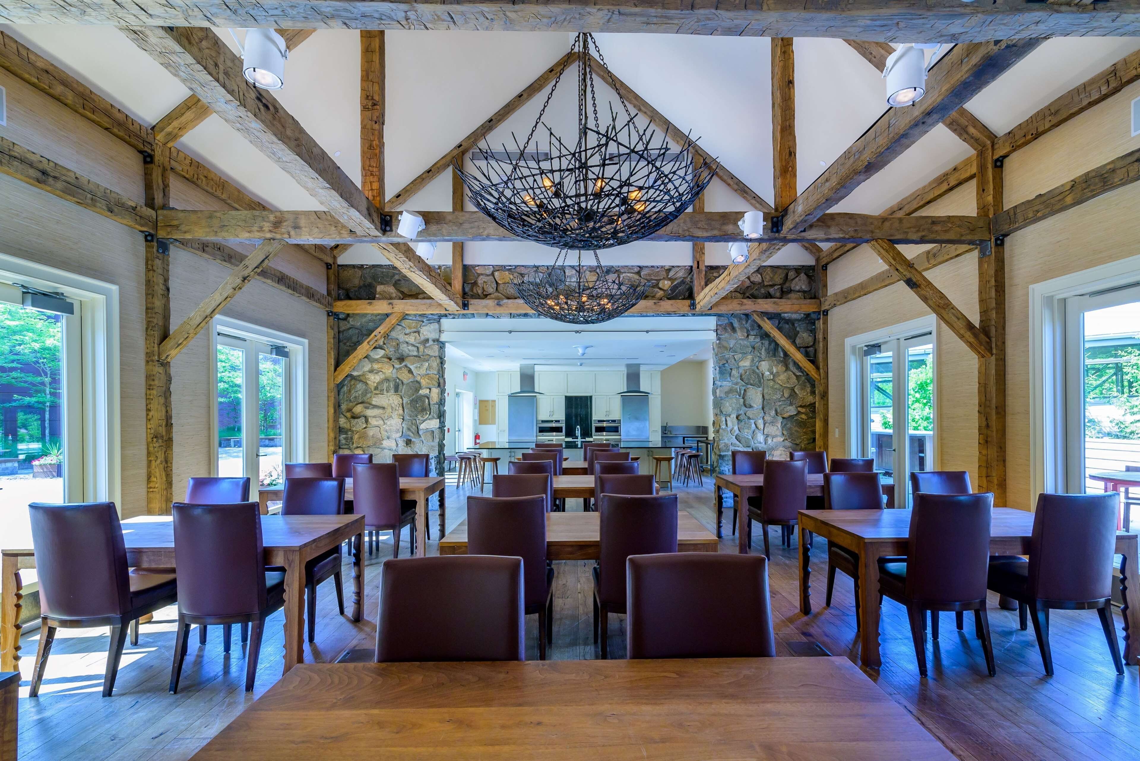 A spacious dining area with wooden beams and stone walls, featuring a central hanging light fixture and multiple tables and chairs.