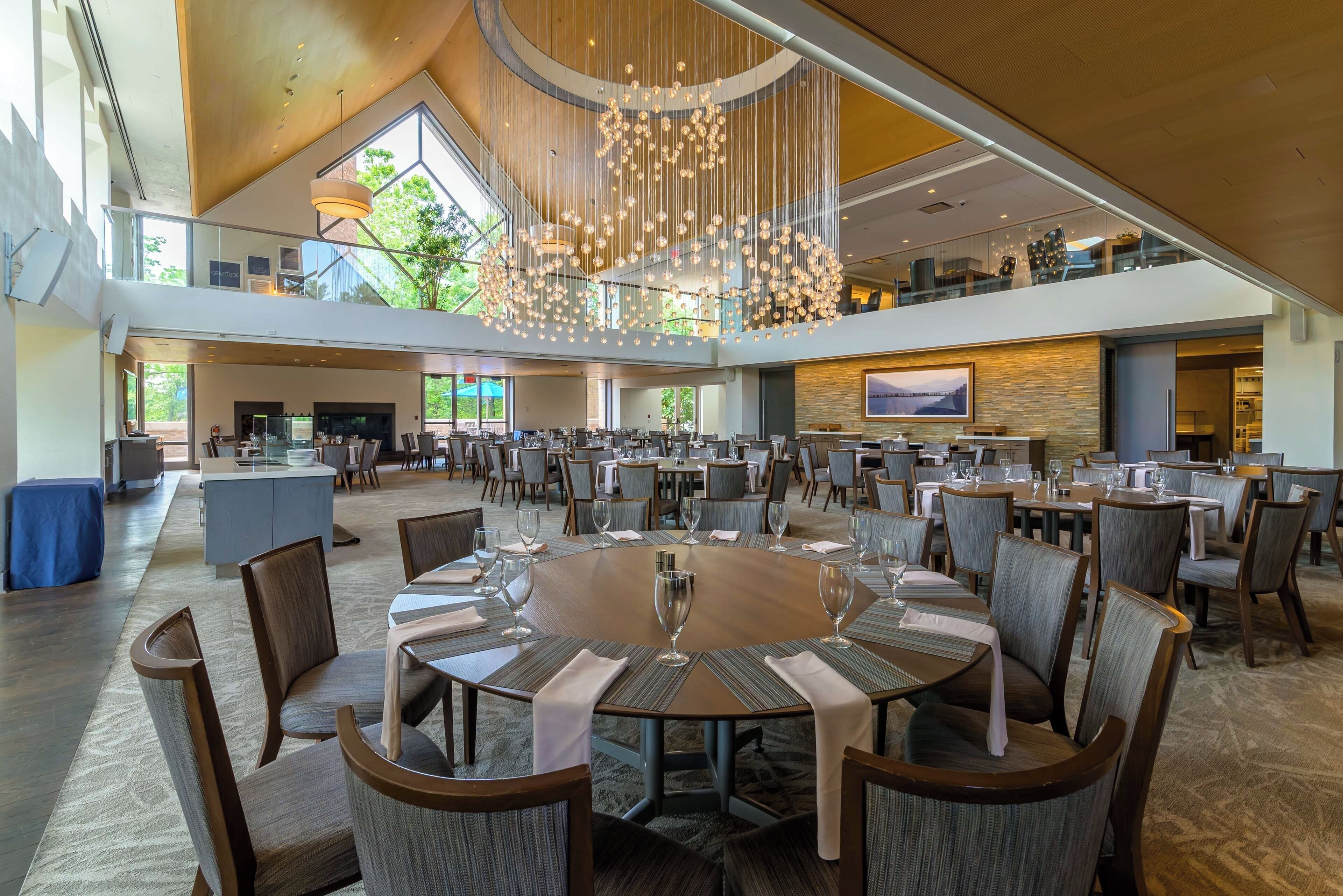 A spacious dining area with multiple tables and chairs, a large chandelier, and a view of the outdoors through large windows.