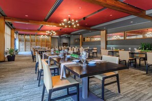 A spacious dining area with multiple tables set for dining, a modern chandelier, and a view of the kitchen area. - Windrose On Hudson (Ossining)