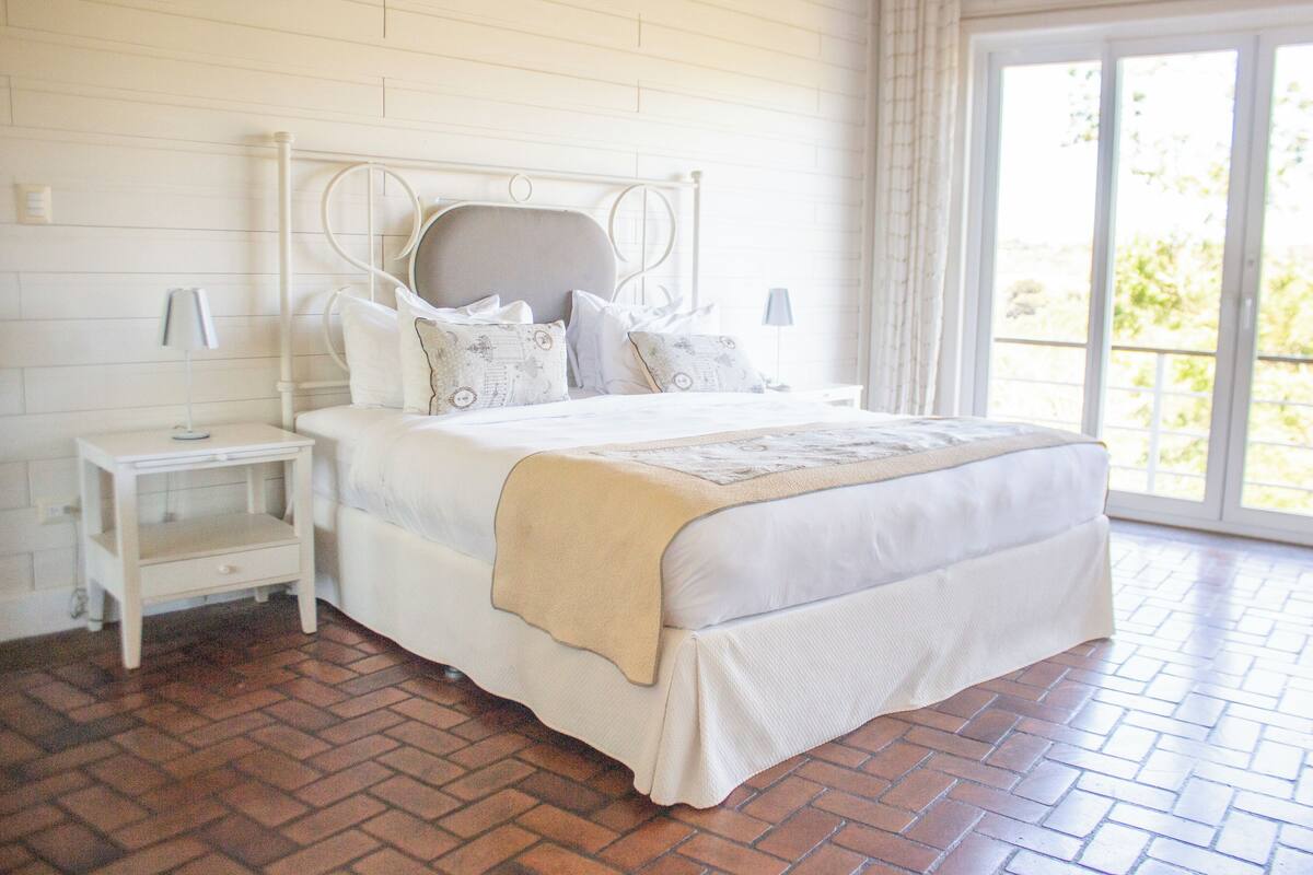 Une chambre avec un lit blanc, une table de chevet, une fenêtre donnant sur un paysage agréable et un plancher en briques.