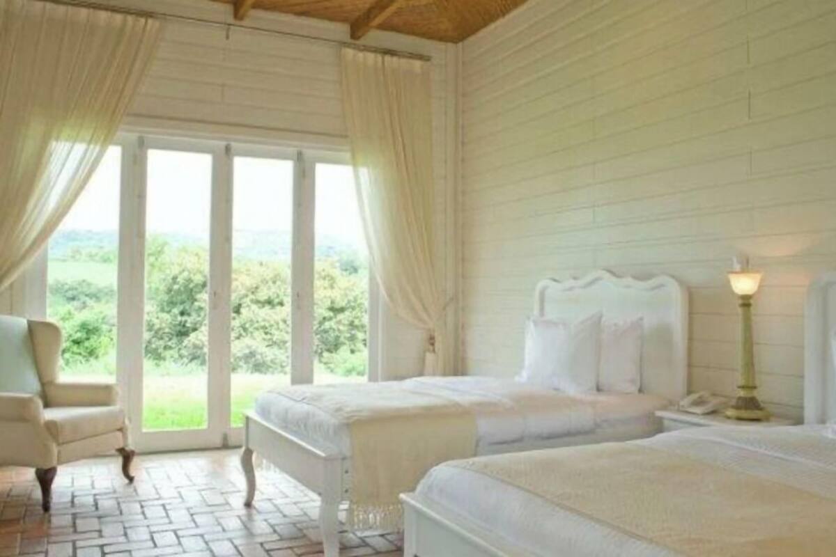 Une chambre avec deux lits, une chaise, une lampe et une vue sur la verdure grâce à de grandes fenêtres.