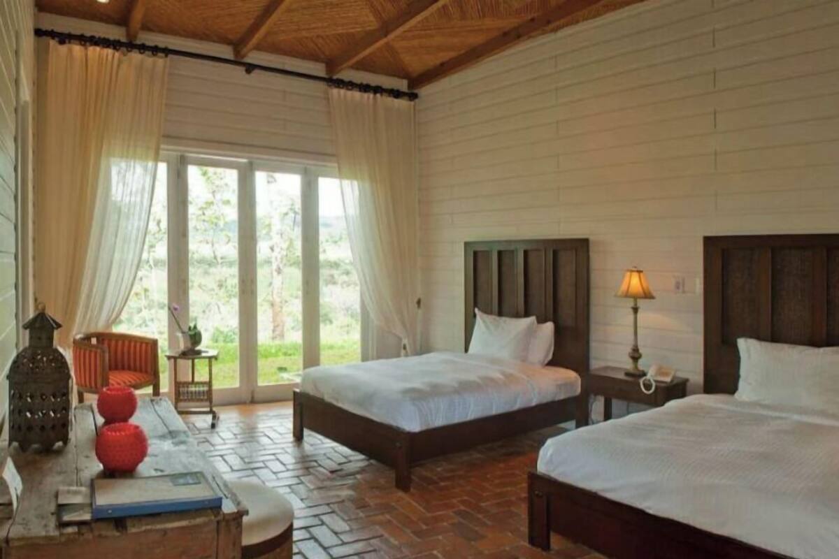 Une chambre d’hôtel avec deux lits, un plancher en bois, une grande fenêtre avec des rideaux, une petite table avec une lampe et une chaise.