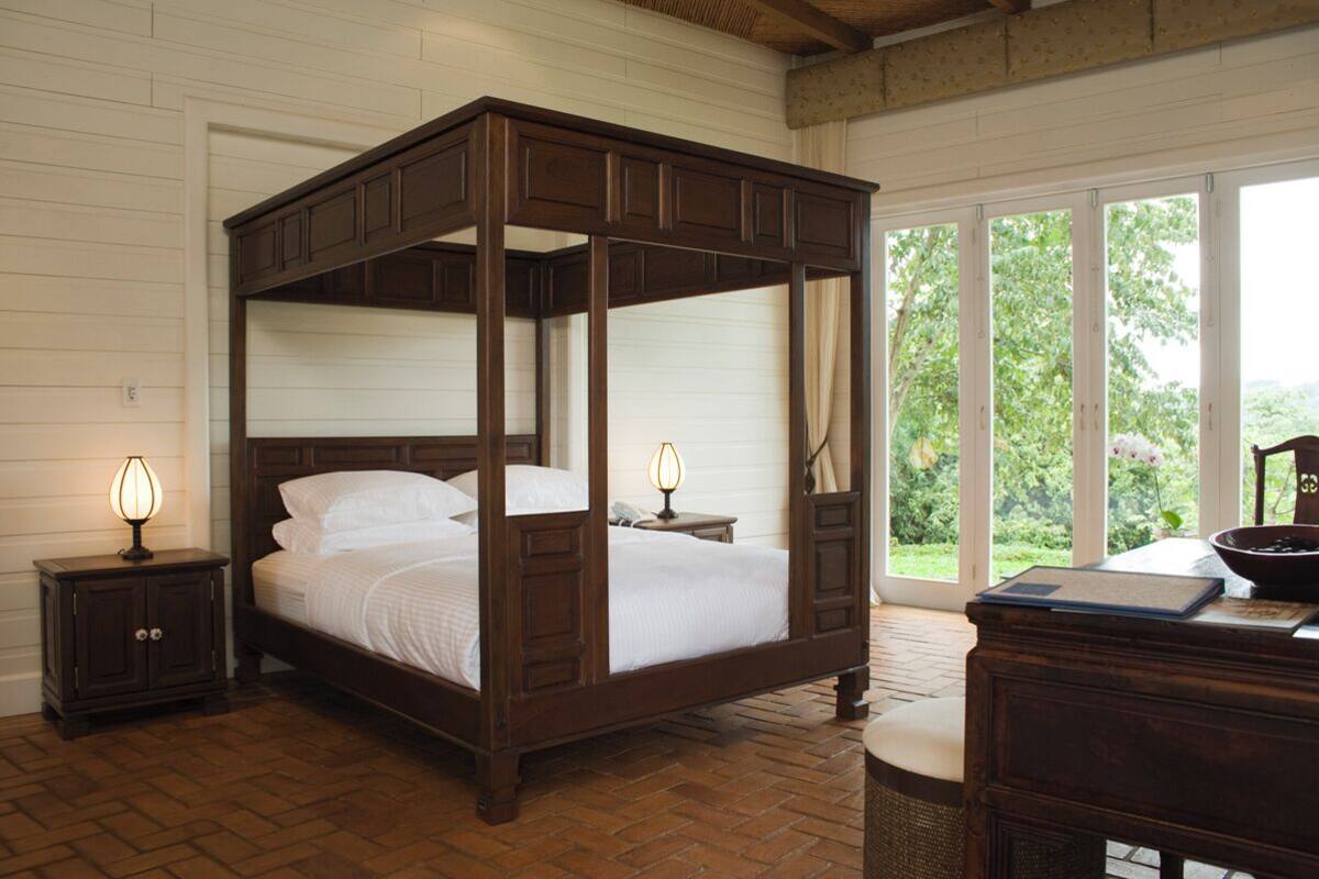 Un lit à baldaquin avec une structure en bois, deux tables de chevet avec des lampes, un bureau avec un bol, et une vue sur la verdure à travers de grandes fenêtres.