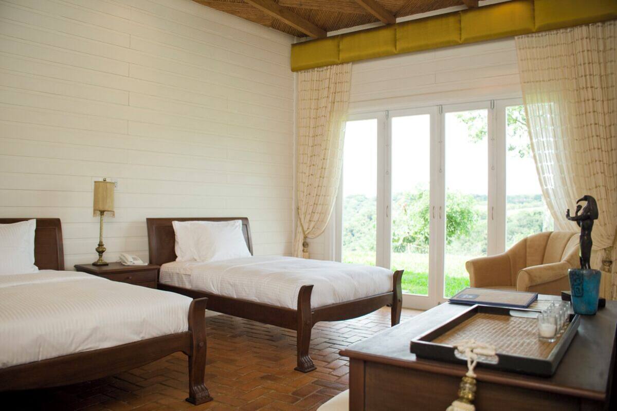 Une chambre avec deux lits, un plancher en bois, une fenêtre avec des rideaux, une chaise et un bureau avec une lampe et une statue.