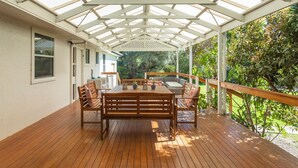 Un patio couvert avec une terrasse en bois, une table centrale entourée de chaises et une vue sur un jardin.