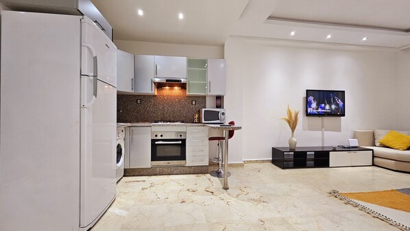 Een moderne keuken met een inbouwoven en magnetron, een eethoek met een klein tafeltje en stoelen, en een woonkamer met een bank en een flatscreen-tv.