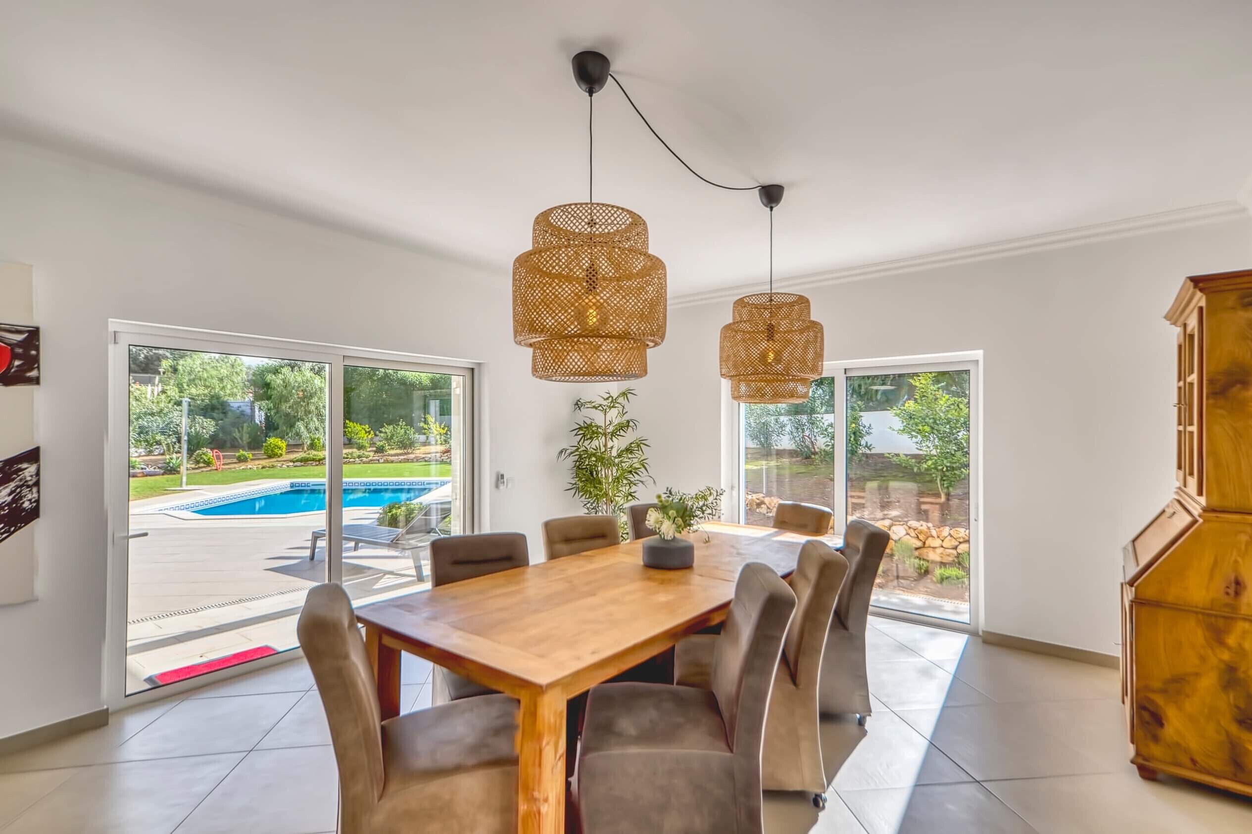 A spacious dining area with a wooden table and chairs, large windows with a view of a swimming pool and garden, and two hanging wicker pendant lights.