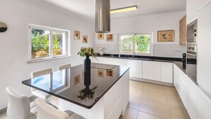 A modern kitchen with white cabinetry, a central island with seating, a stainless steel range hood, and a view of greenery outside. - Villa Larissa, luxury private villa, pool (Portimão)