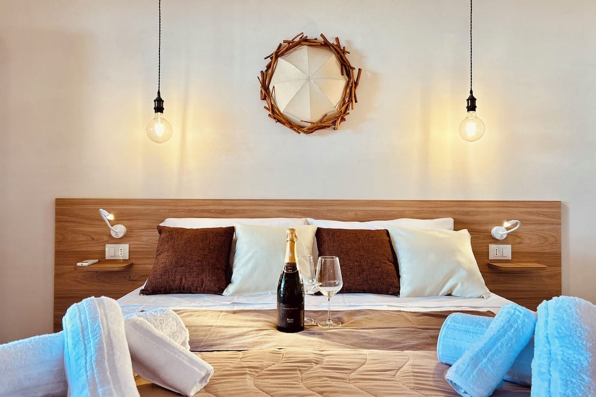 Una camera d'hotel con una testiera in legno, una ghirlanda decorativa, lampade sui comodini, una bottiglia di champagne e asciugamani sul letto.