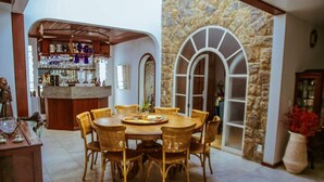 A dining area with a round table and wicker chairs, a bar with shelves of bottles and glasses, and a stone wall with arched windows. - MANSÃO RESIDENCE HOSPEDARIA E EVENTOS (Vassouras)