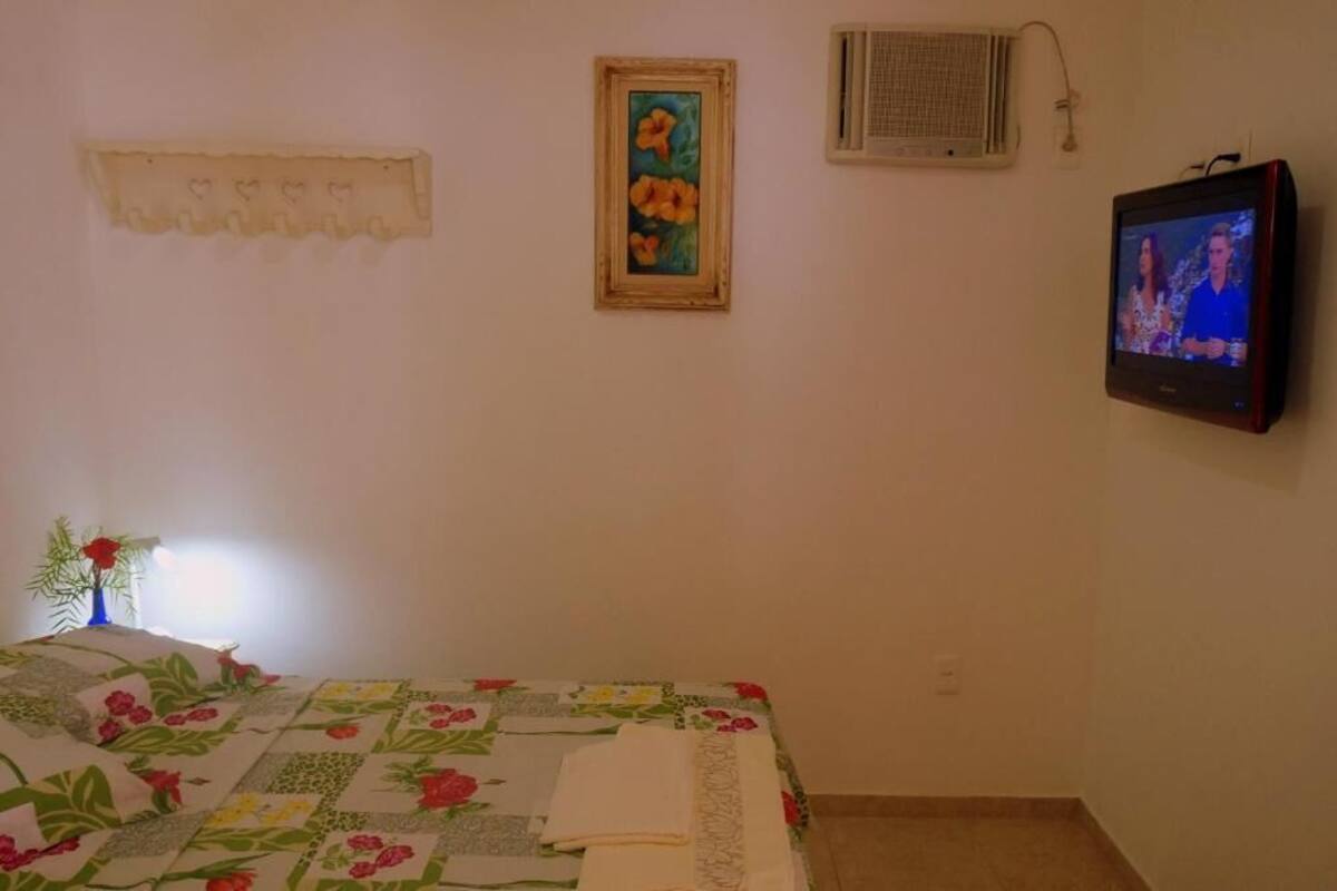 A bedroom with a bed, a wall-mounted TV, a framed painting, and a small window with an air conditioning unit.
