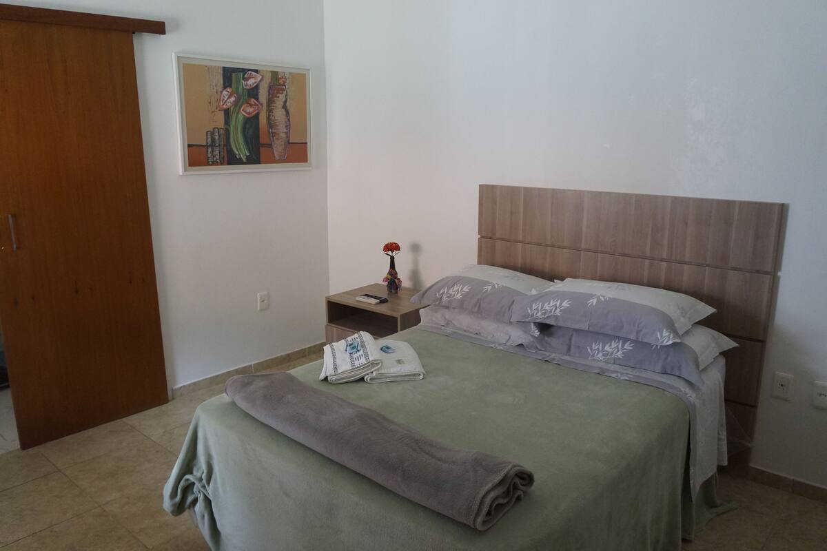 A neatly made bed with a green bedspread and a rolled gray towel, a wooden headboard, a nightstand with a vase of flowers, and a painting on the wall.