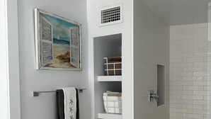 A bathroom with a white tiled shower, a towel rack with a towel, a shelf unit with storage baskets, and a painting of a beach scene. - Steps from the Beach, Surrounded by Serenity – Your Coastal Getaway Awaits (Ormond Beach)