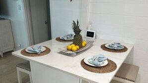 A white dining table with woven placemats, a pineapple, lemons, and a small TV on the table. - Steps from the Beach, Surrounded by Serenity – Your Coastal Getaway Awaits (Ormond Beach)