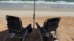 Two beach chairs under a striped umbrella on a sandy beach with the ocean in the background. - Steps from the Beach, Surrounded by Serenity – Your Coastal Getaway Awaits (Ormond Beach)
