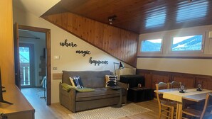 Ein gemütliches Wohnzimmer mit Holzbalkendecke, einer Couch mit Kissen, einem kleinen Tisch und Blick auf Berge durch die Fenster.
