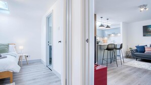 A modern apartment with a wooden floor, a kitchen with bar stools, a dining area, and a living room with a sofa and a painting. - Sandy Steps - 1 Bedroom Apartment - Tenby (Tenby)