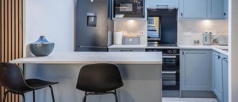 A modern kitchen with a central island, two bar stools, a refrigerator, a microwave, an oven, and a coffee maker.