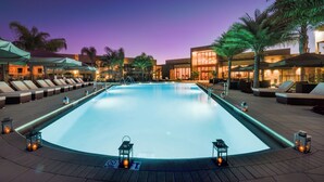 A swimming pool with lounge chairs and umbrellas, surrounded by palm trees and buildings.
