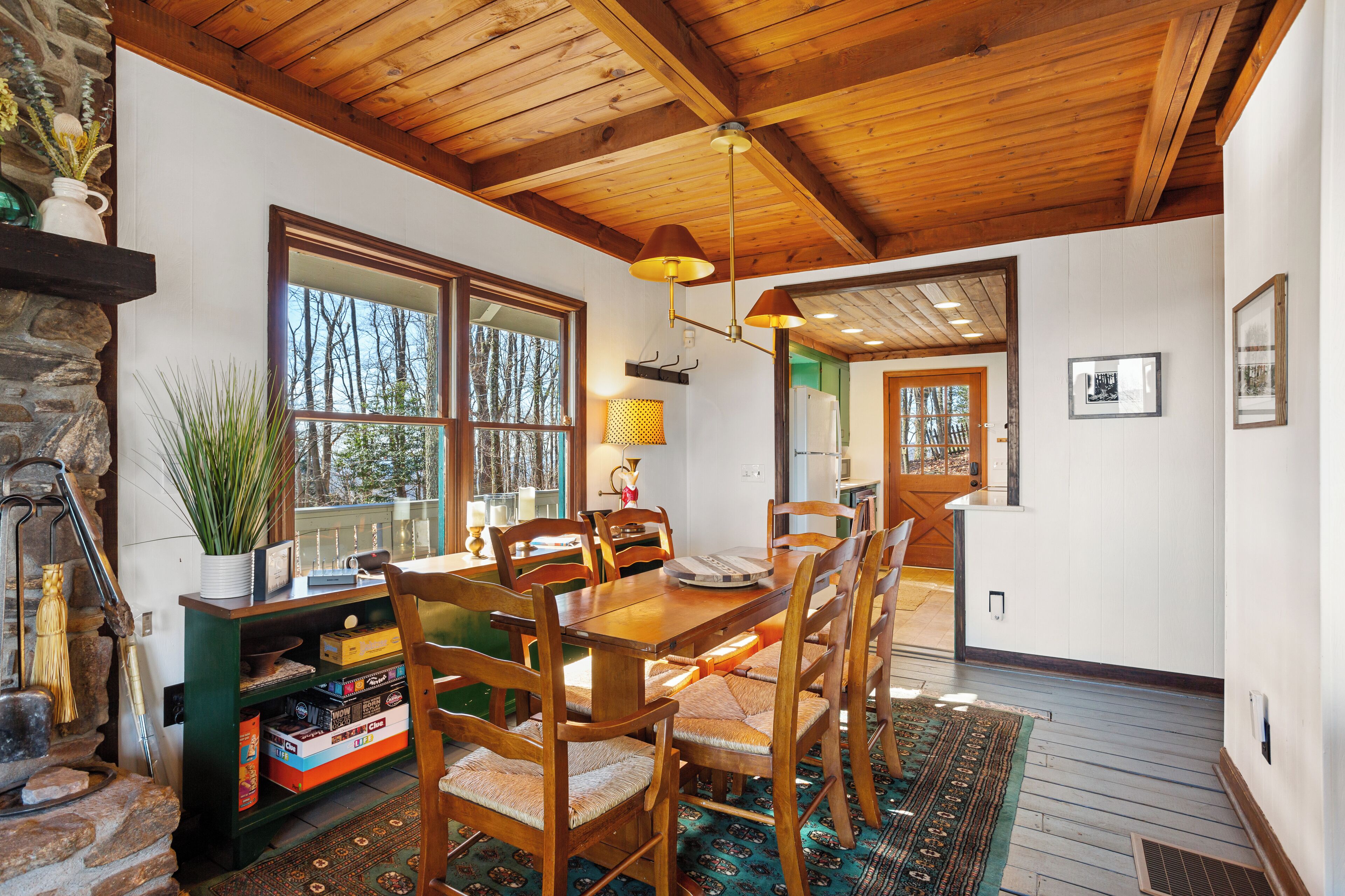 A wooden dining table with chairs, a stone fireplace, and a large window with a view of trees.