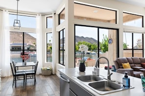 A modern kitchen with large windows, a dining area, and a living space with a sofa and cushions.