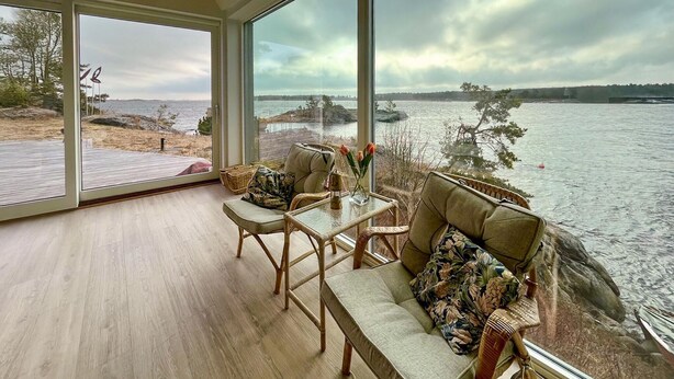 Una habitación con un ventanal grande con vistas a un lago, amueblada con sillas de mimbre y un sofá, y una mesita con un jarrón de flores.