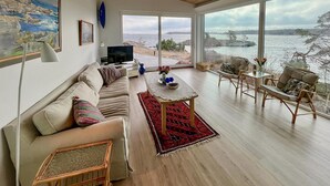 Una sala de estar moderna con un gran ventanal con vistas a un cuerpo de agua, un sofá, una mesa de centro y un comedor con sillas.
