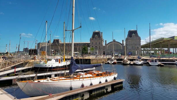 Un port de plaisance avec des bateaux amarrés, un grand bâtiment doté d’une tour horlogée et un espace abrité où sont entreposés des vélos.