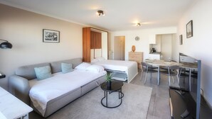 Une pièce moderne et bien éclairée, comprenant un canapé, un lit, une petite table avec une plante, et un coin repas avec des chaises et une table.