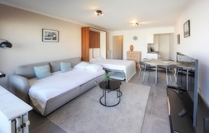 Een moderne, goed verlichte kamer met een bank, een bed, een kleine tafel met een plant en een zithoek met stoelen en een tafel.