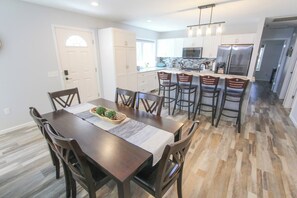 A modern kitchen with white cabinets, a central island, and a dining area with a wooden table and chairs. - A 3-bedroom lake-side cottage at Sunnyside with a fenced yard, sleeps up to 6. (Greenbush)