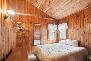 A wooden cabin room with a bed, a mirror, a hanging light fixture, and two windows with curtains. - Book multiple lakefront cottages at Sunnyside, from cozy studios to a 4BR home. (Greenbush)