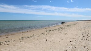 Una playa extensa de arena clara con aguas tranquilas, cielo despejado y tierra a lo lejos.