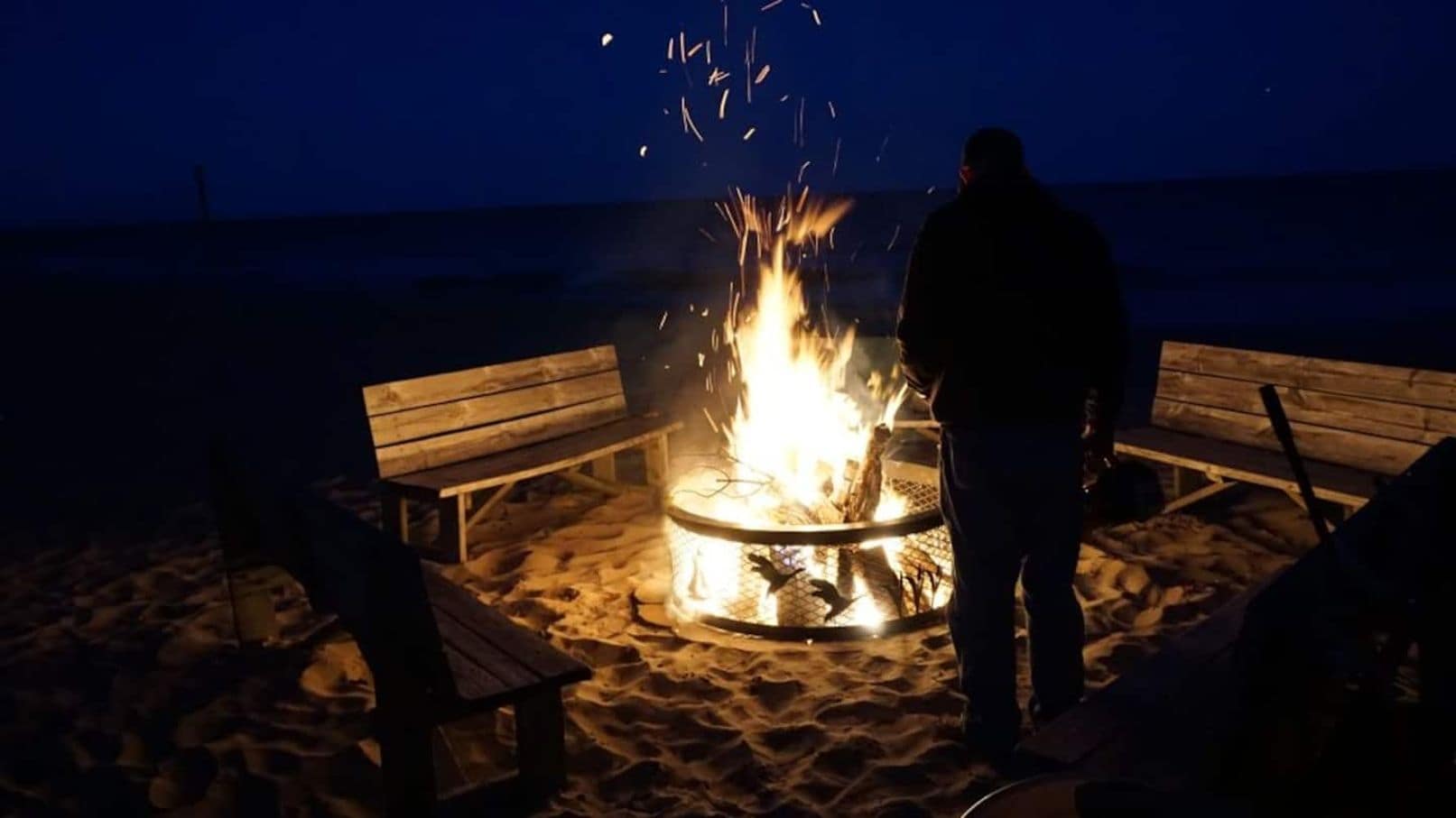Une personne se tient près d'un foyer sur une plage de sable, avec des bancs en bois à proximité.