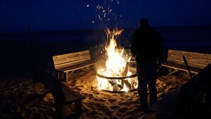Eine Person steht an einem Lagerfeuer an einem sandigen Strand, mit Holzbanken.
