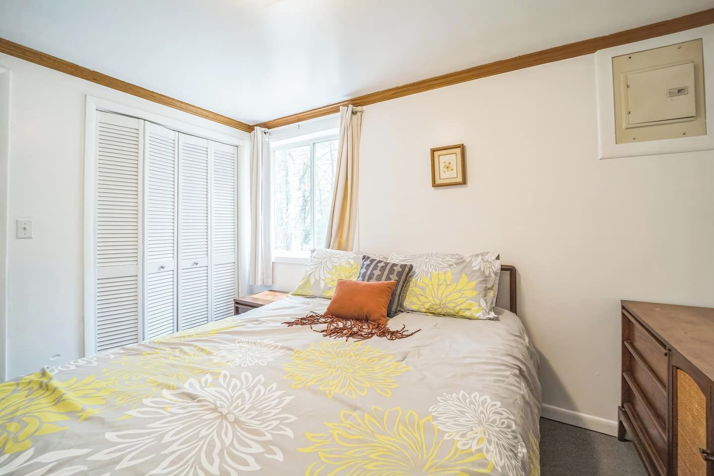 Un dormitorio con cama, ventana con cortinas, cabecero de madera, mesita de noche e interruptor de luz en la pared.