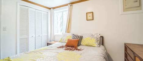A bedroom with a bed, a window with curtains, a wooden headboard, a bedside table, and a wall-mounted light switch.